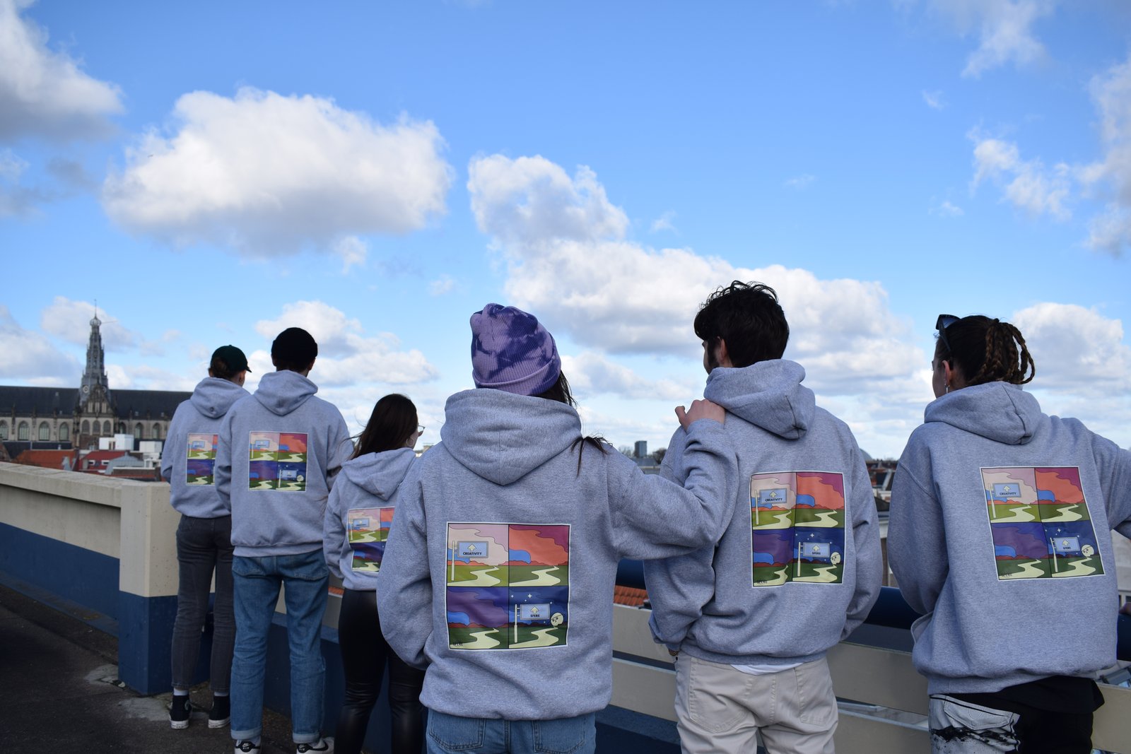Group in grey SFERE hoodies with printed back artwork, looking out over the rooftops
