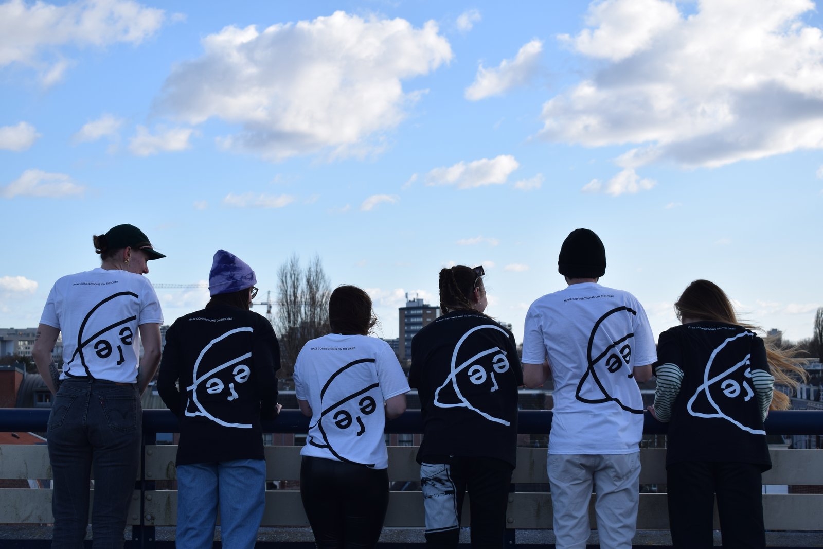 Six models in black and white SFERE t-shirts leaning on a railing, backs to camera